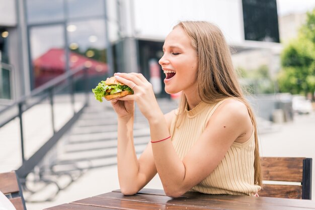 Mooie jonge vrouw die hamburger buiten op straat eet