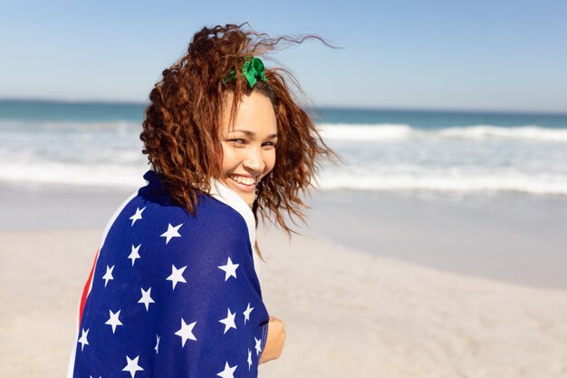 Mooie jonge vrouw die in Amerikaanse vlag wordt verpakt die camera op strand in de zonneschijn bekijkt