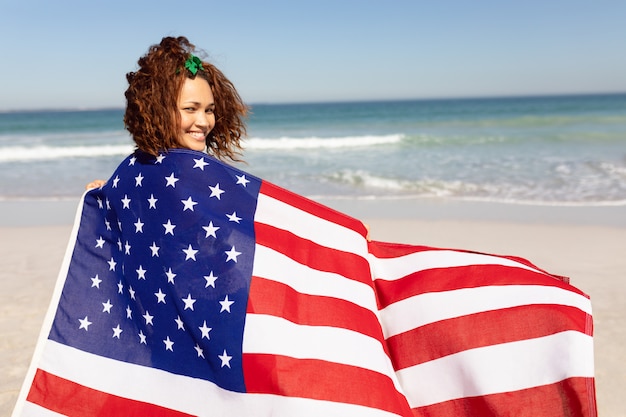 Mooie jonge vrouw die in Amerikaanse vlag wordt verpakt die camera op strand in de zonneschijn bekijkt