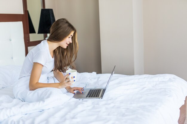 Mooie jonge vrouw die koffie drinkt en op internet surft op haar laptop terwijl ze in bed zit en naar de camera glimlacht