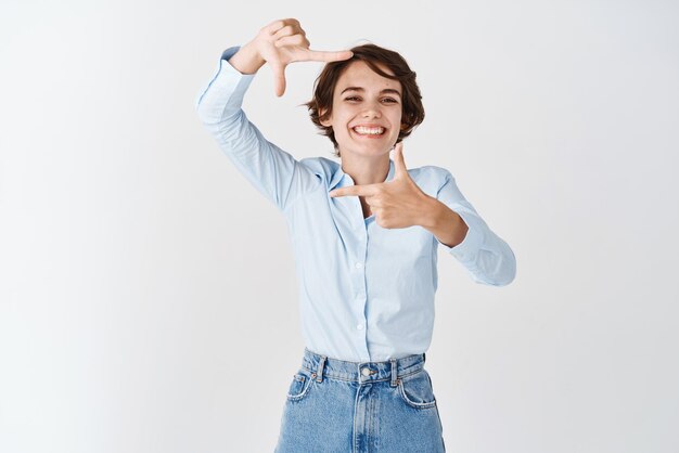 Mooie jonge vrouw die lacht met het gebaar van handframes en doorkijkt naar de camera die een moment vastlegt dat op een witte achtergrond staat