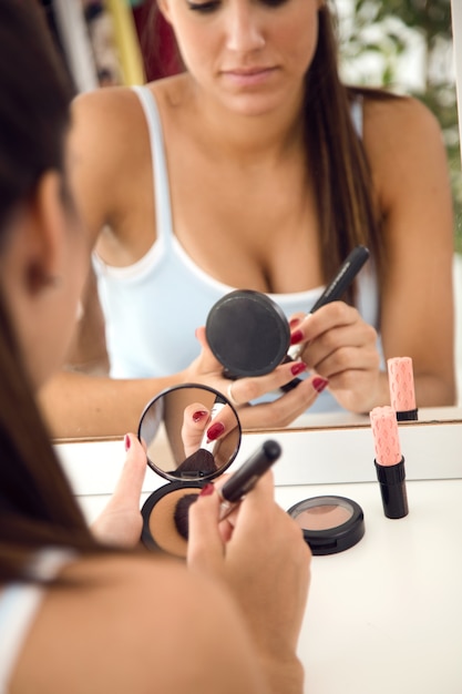 Mooie jonge vrouw die make-up dichtbij spiegel thuis maakt.