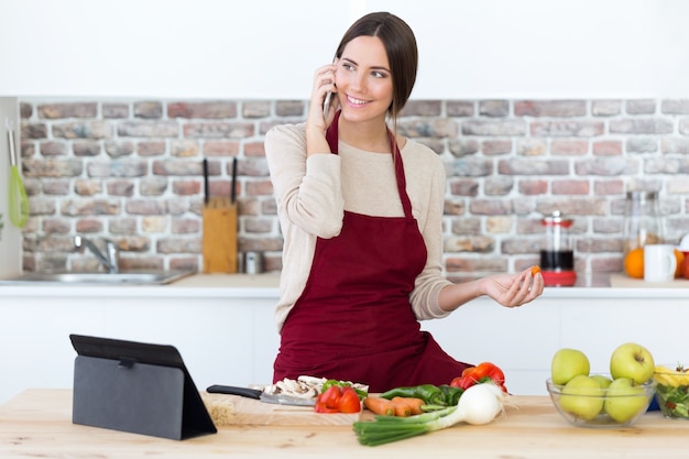 Mooie jonge vrouw die mobiele telefoon met behulp van terwijl het koken in de keuken.