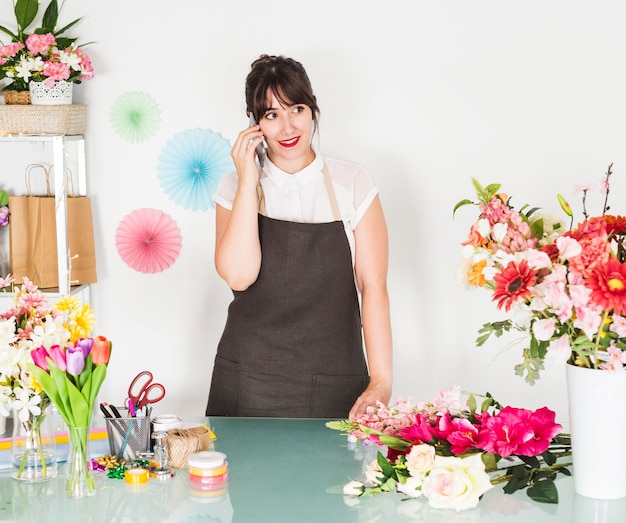 Mooie jonge vrouw die op cellphone met bloemen op bureau spreekt