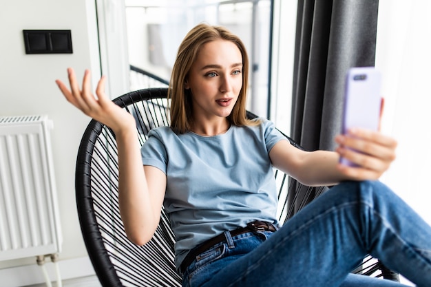 Mooie jonge vrouw die selfie maakt met haar smartphone en glimlacht terwijl ze thuis in een grote stoel zit