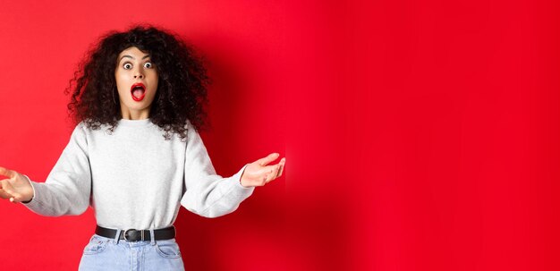 Gratis foto mooie jonge vrouw die vol ontzag staat, laat haar kaak vallen en kijkt onder de indruk naar de camera, spreidt haar handen zijwaarts