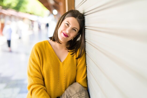 Mooie jonge vrouw glimlachend zelfverzekerd en vrolijk leunend op een houten muur die op een zonnige dag door de straat van de stad loopt