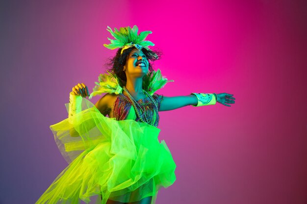 Mooie jonge vrouw in carnaval, stijlvol maskeradekostuum met veren die op gradiëntachtergrond dansen in neonlicht.