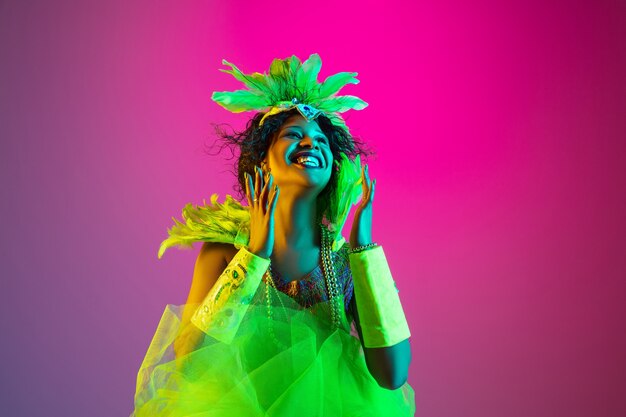 Mooie jonge vrouw in Carnaval, stijlvol maskeradekostuum met veren die op gradiëntmuur dansen in neonlicht