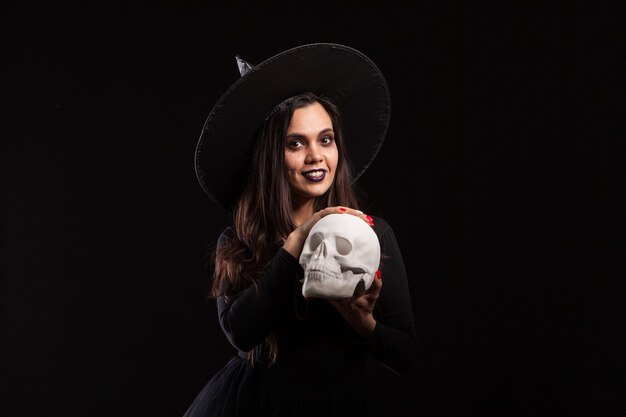 Mooie jonge vrouw in een heksenkostuum voor halloween-feest dat kwade hekserij doet. Portret van een vrouw die magie doet op een menselijke schedel.