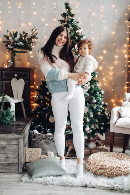 Mooie jonge vrouw in pluizige trui glimlachend terwijl het kind vasthoudt en in de buurt van de kerstboom staat