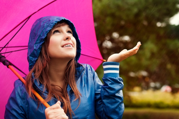 Mooie jonge vrouw in regenjas met paraplu die op regen controleert