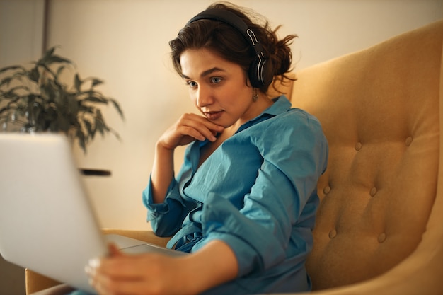 Mooie jonge vrouw met behulp van draadloze headset tijdens het kijken naar webinar op sociale media marketing, met gerichte blik. Mooi meisje leren online zittend op de bank met laptop, hand op kin te houden