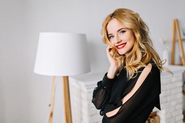 Mooie jonge vrouw met blond golvend haar poseren in gezellige kamer met witte meubels, genieten van een goede dag thuis. Het dragen van elegante zwarte blouse, lichte dagmake-up met rode lippenstift.