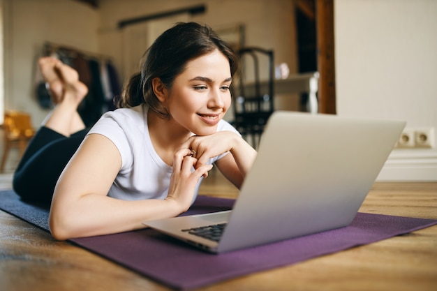 Mooie jonge vrouw met gespierde armen met behulp van laptop voor thuistraining, video cursus online kijken, moet binnenshuis oefenen terwijl sociale afstand nemen, met een geïnsireerde blik.