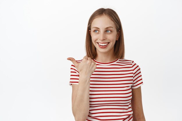 Mooie jonge vrouw met kort blond haar, wijzend en naar links kijkend, promotekst lezend met een tevreden gelukkige glimlach, staande in gestreept t-shirt op witte achtergrond.