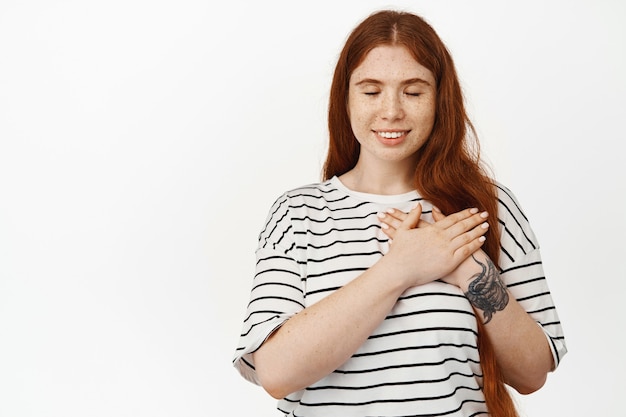 Mooie jonge vrouw met lang rood haar en bleke huid, heeft herinneringen, dagdromen, hand in hand op het hart en glimlachend met gesloten ogen, onthoud iets op wit