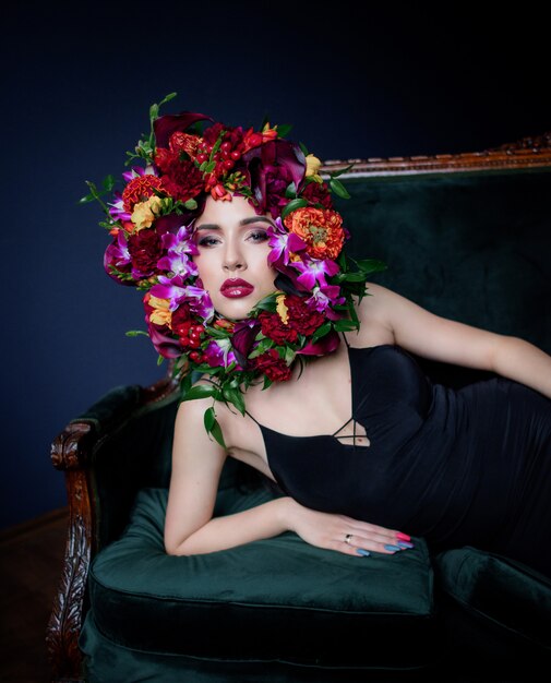 Mooie jonge vrouw met lichte make-up ligt op de groene bank, gezicht omringd met kleurrijke verse bloemen op de donkerblauwe achtergrond