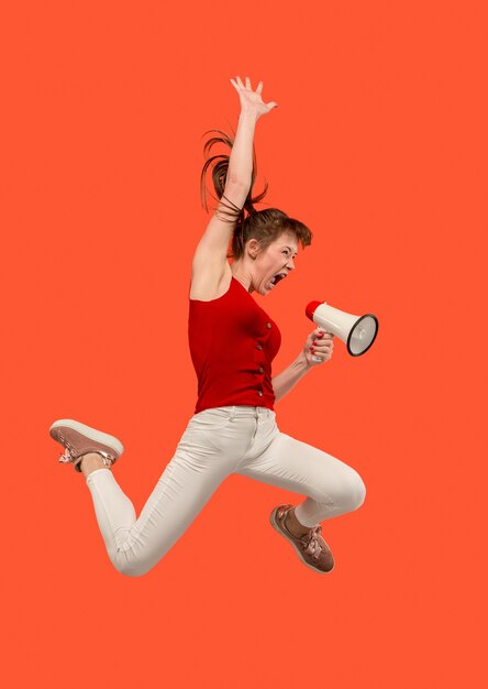 Mooie jonge vrouw springen met megafoon geïsoleerd op rode achtergrond. Runnin meisje in beweging of beweging. Menselijke emoties en gezichtsuitdrukkingen concept