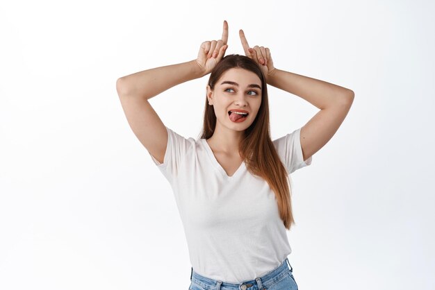 Mooie jonge vrouw toont tong en stier hoorns gebaar opzij kijken naar kopieerruimte met promotionele tekst lezen logo teken staande in tshirt tegen witte achtergrond