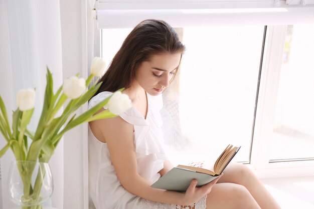 Mooie jonge vrouw zittend op de vensterbank en boek lezen