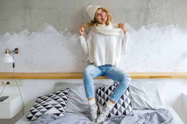 Gratis foto mooie jonge vrouwenzitting in slaapkamer tegen muur die witte sweater draagt