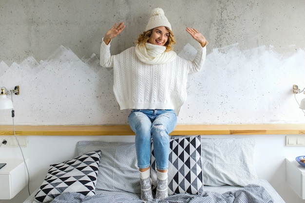 Mooie jonge vrouwenzitting in slaapkamer tegen muur die witte sweater draagt