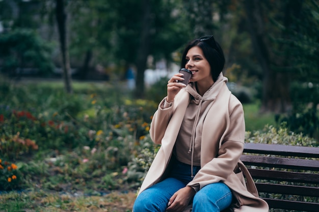 Mooie jonge vrouwenzitting op een bank het drinken koffie die in park genieten van