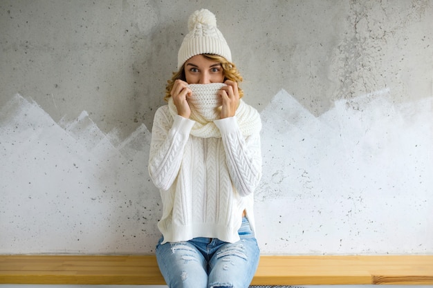 Gratis foto mooie jonge vrouwenzitting tegen muur die witte sweater, gebreide muts en sjaal draagt