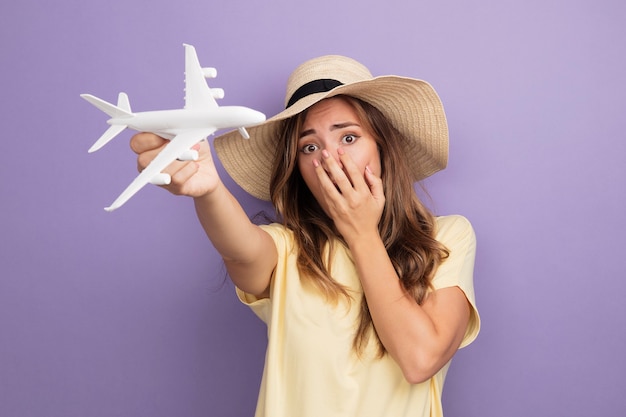 Mooie jongedame in beige t-shirt en zomerhoed met speelgoedvliegtuig kijkend naar camera die geschokt is over mond met hand die over paarse achtergrond staat