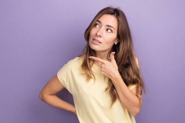 Mooie jongedame in beige t-shirt opzij kijkend met een glimlach op een slim gezicht denkend over een paarse achtergrond