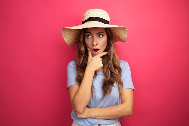 Mooie jongedame in blauw t-shirt en zomerhoed die opzij kijkt verbaasd over roze achtergrond