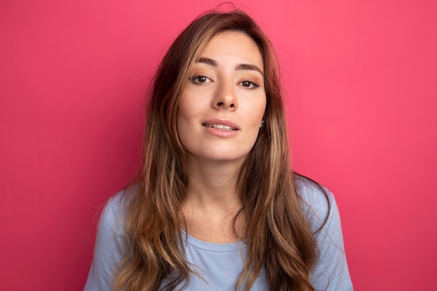 Mooie jongedame in blauw t-shirt kijkend naar camera glimlachend zelfverzekerd over roze achtergrond