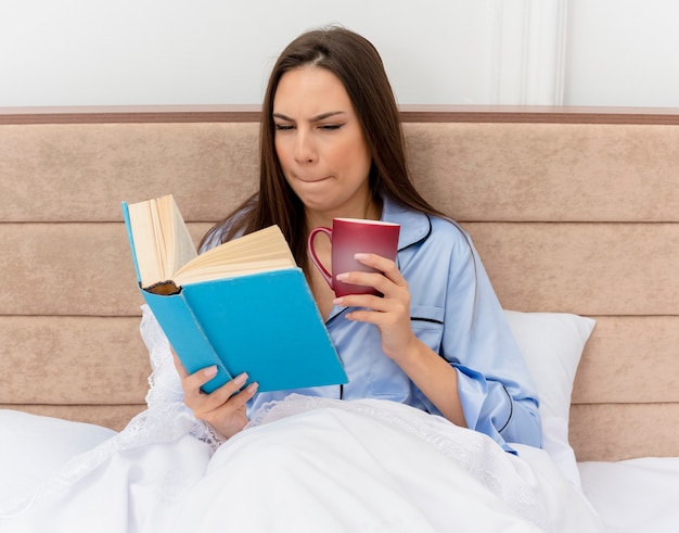 Mooie jongedame in blauwe pyjama liggend op bed met boek en kopje koffie op zoek met sceptische uitdrukking in slaapkamer interieur op lichte achtergrond