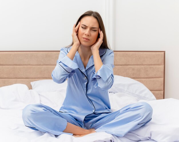 Mooie jongedame in blauwe pyjama zittend op bed tempels aanraken op zoek onwel met sterke hoofdpijn in slaapkamer interieur op lichte achtergrond