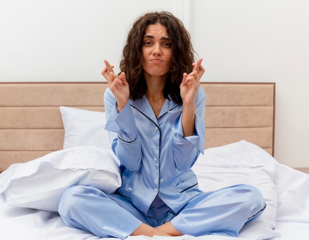 Mooie jongedame in blauwe pyjama zittend op bed wenselijke wens kruising vingers kijken ontevreden in slaapkamer interieur op lichte achtergrond maken