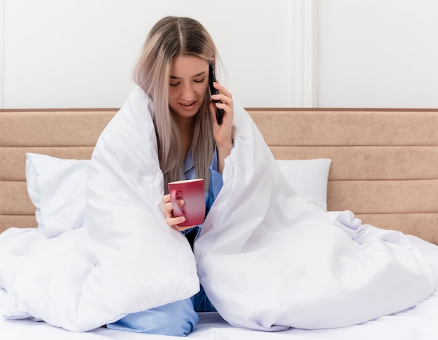 Mooie jongedame in blauwe pyjama zittend op bed wikkelen in deken met kopje koffie praten op mobiele telefoon in slaapkamer interieur