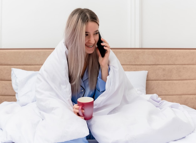 Mooie jongedame in blauwe pyjama zittend op bed wikkelen in deken met kopje koffie praten op mobiele telefoon in slaapkamer interieur