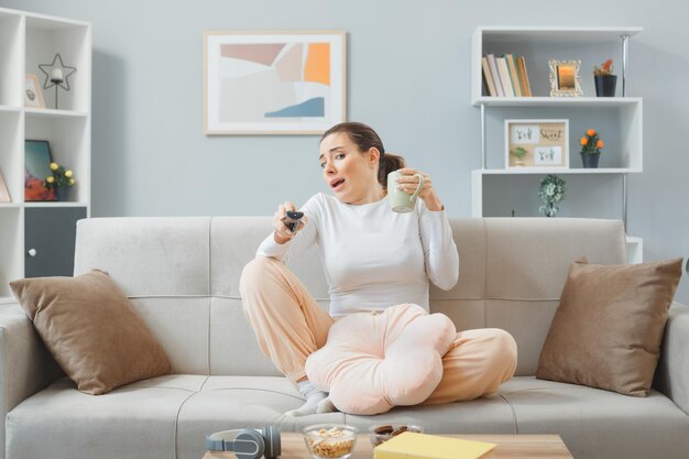 Mooie jongedame in casual kleding zittend op een bank in het interieur met afstandsbediening en een kopje thee die er bang en bezorgd uitziet en een weekend thuis doorbrengt