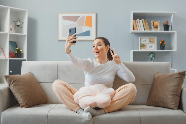 Mooie jongedame in casual kleding zittend op een bank in het interieur van het huis met smartphone met videogesprek of een selfie maken gelukkig en positief tonen vsign terwijl u thuis ontspant
