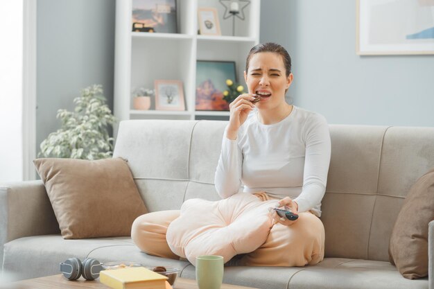 Mooie jongedame in casual kleding zittend op een bank thuis interieur eten koekje met afstandsbediening kijken verward en ontevreden maken wrange mond weekend doorbrengen thuis