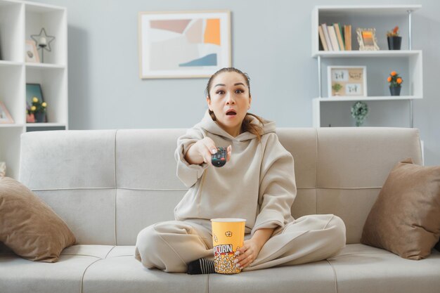 Mooie jongedame in huiskleding zittend op een bank in het interieur met een emmer popcorn die op afstand televisie kijkt, verrast en verbaasd om tijd thuis door te brengen