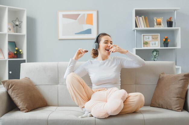 Mooie jongedame in huiskleding zittend op een bank in het interieur met koptelefoon die koekjes eet, gelukkig en positief ontspannen, plezier hebbend weekend thuis doorbrengen