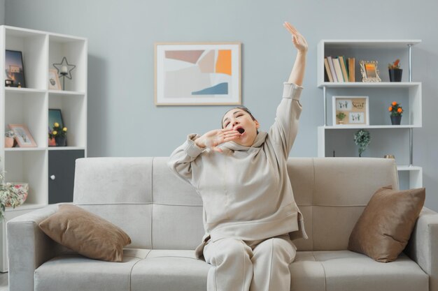 Mooie jongedame in huiskleding zittend op een bank in het interieur van het huis en ziet er moe en verveeld uit, geeuwen en uitrekkend tijd thuis doorbrengend