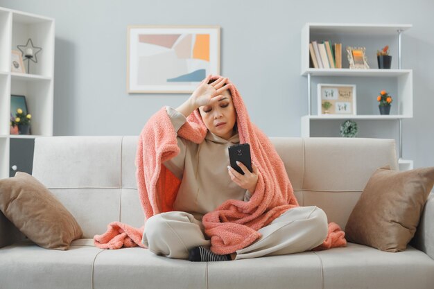 Mooie jongedame in huiskleding zittend op een bank onder deken in het interieur met smartphone die er onwel uitziet met hoofdpijn en temperatuur