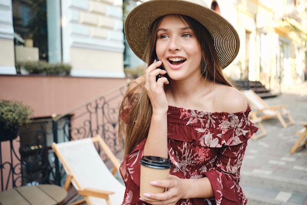 Mooie jongedame in jurk en strohoed met kopje koffie in de hand om te gaan praten op mobiel terwijl ze gelukkig opzij kijkt op gezellige straat in de stad