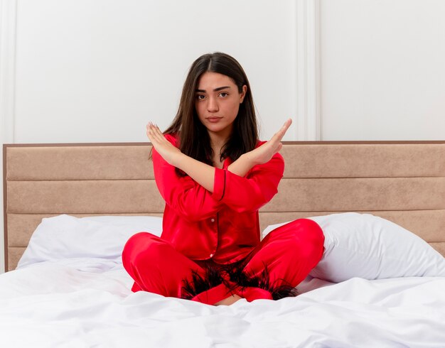 Mooie jongedame in rode pyjama's die op bed zit en een stopgebaar maakt en handen kruist met een serieus gezicht in het interieur van de slaapkamer