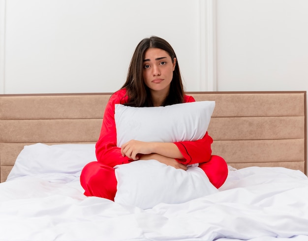 Mooie jongedame in rode pyjama zittend in bed knuffelend kussen ongelukkig met droevige uitdrukking in slaapkamerinterieur