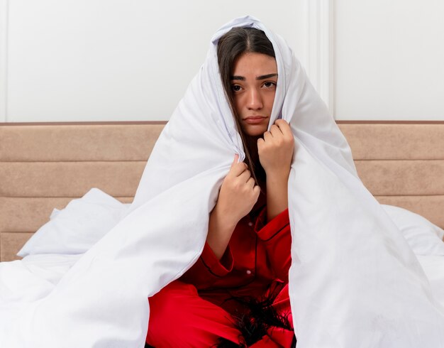 Mooie jongedame in rode pyjama zittend op bed inwikkeling in deken camera kijken met droevige uitdrukking in slaapkamer interieur op lichte achtergrond