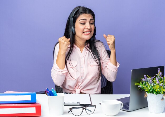 Mooie jongedame in vrijetijdskleding met een hoofdtelefoon met microfoon balde vuisten boos en teleurgesteld zittend aan tafel met laptop op blauw